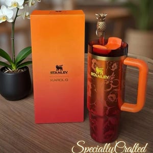 May include: A red and orange Stanley Karol G travel mug with a floral pattern, a matching box, and a pineapple-shaped lid. The words "Customisable" and "Specially Crafted" are also visible.