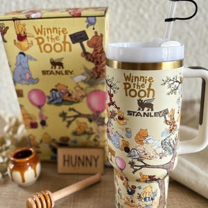 May include: A Winnie the Pooh themed Stanley tumbler with a straw and bee topper, a matching box, and a honey dipper. The tumbler and box feature illustrations of Pooh and friends on a cream background. A small honey pot and a wooden sign that says "Hunny" are also present.