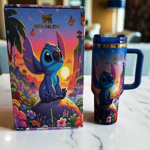 May include: A blue and purple Stanley travel mug and matching box featuring a colorful illustration of Stitch from Lilo & Stitch. The mug has a blue handle and lid. The box and mug have the Stanley logo.