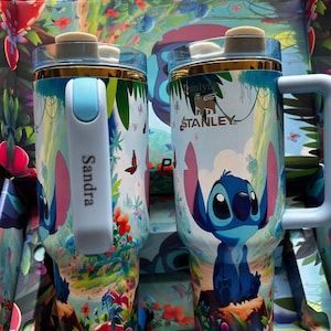 May include: Two Stanley tumblers featuring a colorful design with the character Stitch. The tumblers have a light blue handle and lid, with a gold rim. The background includes a jungle scene with flowers and butterflies. The text "Sandra" is printed on one of the handles.