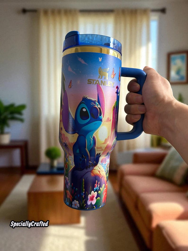 May include: A blue Stanley tumbler featuring a colorful design of the character Stitch from Lilo & Stitch. The tumbler has a blue handle and lid, with a gold band at the top. The design includes Stitch sitting on a rock with flowers and a sunset background.