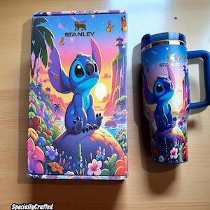 May include: A Stanley brand tumbler and box set featuring a blue cartoon character design. The box and tumbler have a matching design with the character sitting on a rock with a tropical background. The tumbler has a blue handle and lid.