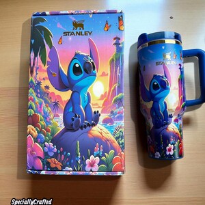 May include: A blue Stanley tumbler and matching box featuring a cartoon character design. The tumbler has a blue handle and lid, with the character image repeated on both items. The box and tumbler have a colorful tropical landscape background.