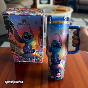 May include: A blue Stanley tumbler and matching box featuring a colorful illustration of a cartoon character. The tumbler has a blue handle and lid, with a gold rim. The box and tumbler have the Stanley logo.
