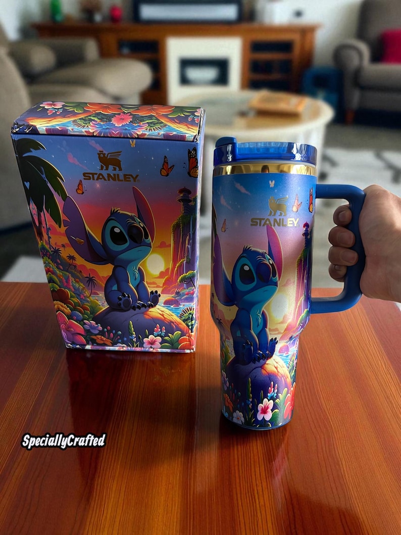 May include: A blue Stanley tumbler and matching box featuring a colorful illustration of a cartoon character. The tumbler has a blue handle and lid, with a gold rim. The box and tumbler have the Stanley logo.