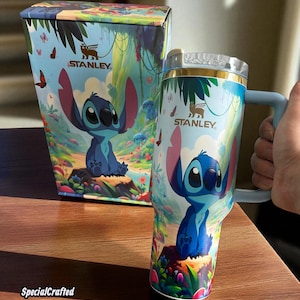 May include: A Stanley tumbler and matching box feature a colorful design with the character Stitch. The tumbler has a blue handle and a clear lid. The box and tumbler have the Stanley logo and the text "SpecialCrafted".
