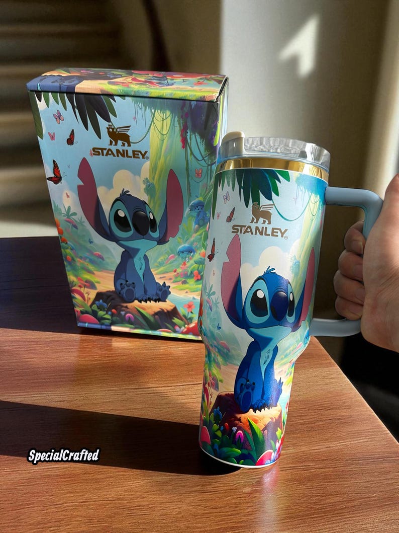 May include: A Stanley tumbler and matching box feature a colorful design with the character Stitch. The tumbler has a blue handle and a clear lid. The box and tumbler have the Stanley logo and the text "SpecialCrafted".