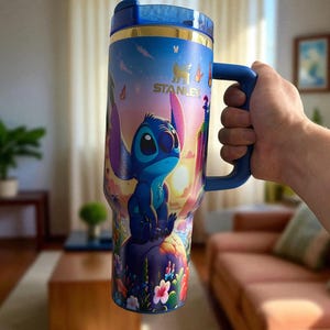 May include: A blue and gold Stanley tumbler featuring the character Stitch from Lilo & Stitch. The tumbler has a blue handle and lid, and a colorful design of Stitch on a tropical island. The tumbler is held by a person.