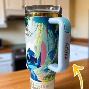 May include: A Stanley Quencher H2.0 tumbler featuring a colorful design of the character Stitch. The tumbler has a light blue handle with the name "Arisha" printed on it. The tumbler is on a wooden surface.