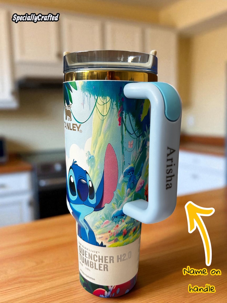 May include: A Stanley Quencher H2.0 tumbler featuring a colorful design of the character Stitch. The tumbler has a light blue handle with the name "Arisha" printed on it. The tumbler is on a wooden surface.