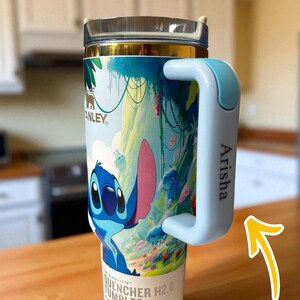 May include: A stainless steel tumbler featuring a colorful illustration of the character Stitch from Lilo & Stitch. The tumbler has a light blue handle with the name "Arisha" printed on it. The tumbler is labeled "Quencher H2.0 Tumbler".