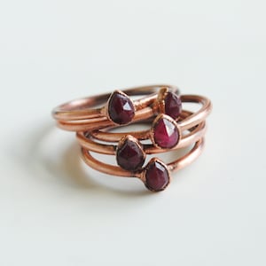 May include: Set of five copper rings with teardrop-shaped red gemstones. The rings are stacked on top of each other.