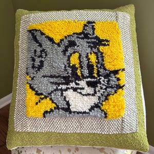 May include: A square decorative pillow featuring a cartoon cat character on a yellow background. The cat is gray, black, and white, with a surprised expression. The pillow has a green border and a textured gray and white frame.