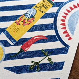 Zoom sur l'illustration. Dessin d'un tube de harissa sur un fond à rayures bleu et blanc. L'illustration est dessinée aux crayons de couleurs. Elle présente également des tomates, piments, ail et coriandre et un plat à motifs, suggérant une ambiance tunisienne. L'illustration est signée de Lucie Leforestier et numérotée, en édition limitée. Le papier est texturé.