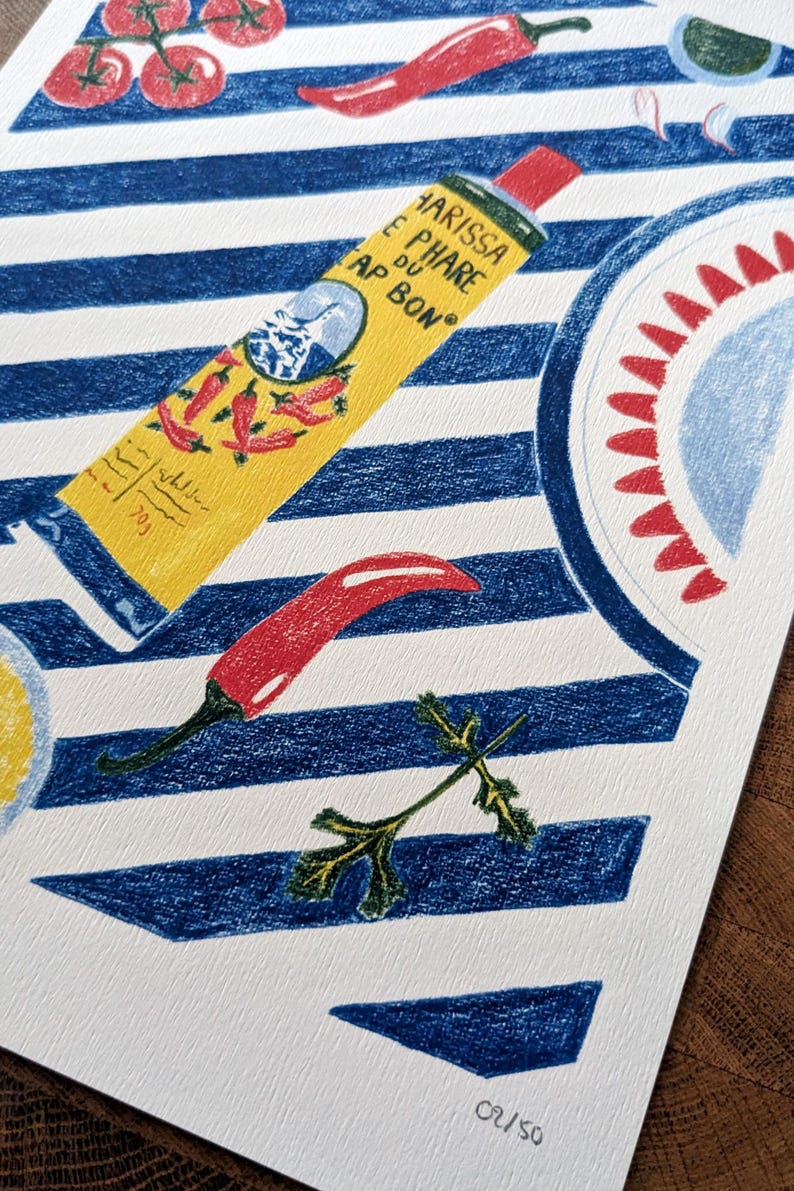Zoom sur l'illustration. Dessin d'un tube de harissa sur un fond à rayures bleu et blanc. L'illustration est dessinée aux crayons de couleurs. Elle présente également des tomates, piments, ail et coriandre et un plat à motifs, suggérant une ambiance tunisienne. L'illustration est signée de Lucie Leforestier et numérotée, en édition limitée. Le papier est texturé.