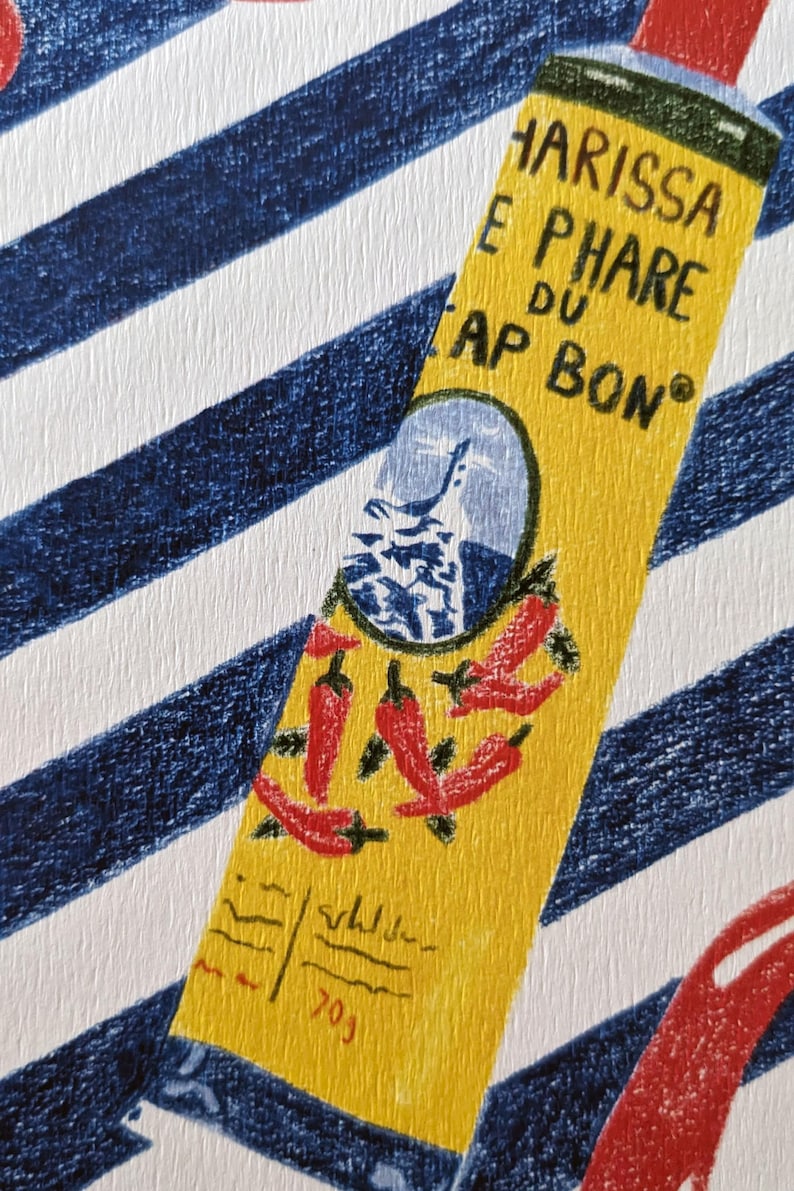 Zoom sur l'illustration. Dessin d'un tube de harissa sur un fond à rayures bleu et blanc. L'illustration est dessinée aux crayons de couleurs. Elle présente également des tomates, piments, ail et coriandre et un plat à motifs, suggérant une ambiance tunisienne. L'illustration est signée de Lucie Leforestier et numérotée, en édition limitée. Le papier est texturé.