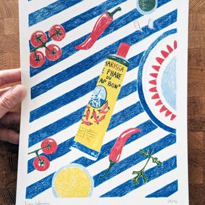 Dessin d'un tube de harissa sur un fond à rayures bleu et blanc. L'illustration est dessinée aux crayons de couleurs. Elle présente également des tomates, piments, ail et coriandre et un plat à motifs, suggérant une ambiance tunisienne. L'illustration est signée de Lucie Leforestier et numérotée, en édition limitée. Une main tient l'illustration.