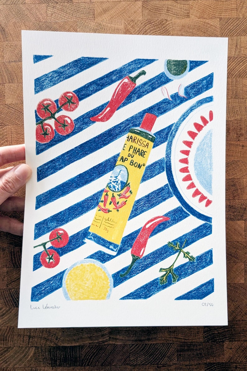 Dessin d'un tube de harissa sur un fond à rayures bleu et blanc. L'illustration est dessinée aux crayons de couleurs. Elle présente également des tomates, piments, ail et coriandre et un plat à motifs, suggérant une ambiance tunisienne. L'illustration est signée de Lucie Leforestier et numérotée, en édition limitée. Une main tient l'illustration.
