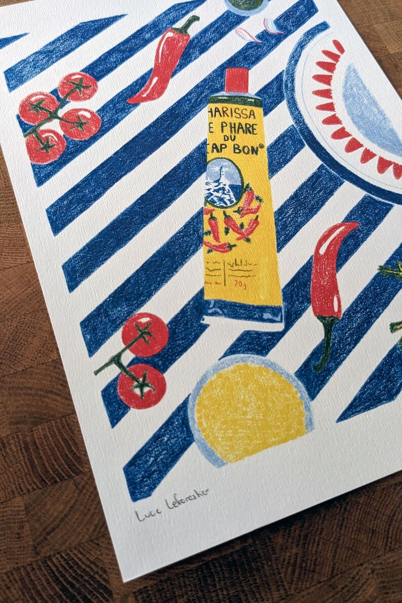 Zoom sur l'illustration. Dessin d'un tube de harissa sur un fond à rayures bleu et blanc. L'illustration est dessinée aux crayons de couleurs. Elle présente également des tomates, piments, ail et coriandre et un plat à motifs, suggérant une ambiance tunisienne. L'illustration est signée de Lucie Leforestier et numérotée, en édition limitée. Le papier est texturé.