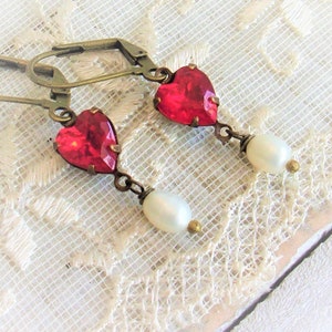 Peut inclure: Boucles d'oreilles de style vintage avec des cristaux rouges en forme de cœur. Chaque boucle d'oreille est ornée d'une perle blanche et de détails en métal couleur laiton antique. Présentées sur un fond de dentelle blanche.
