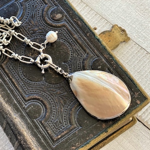 Puede incluir: Un collar de plata con un colgante grande en forma de lágrima. El colgante es de un color claro e iridiscente, y el collar tiene un diseño de eslabones de cadena. Una pequeña perla está unida cerca del cierre. El collar descansa sobre una superficie oscura y texturizada.