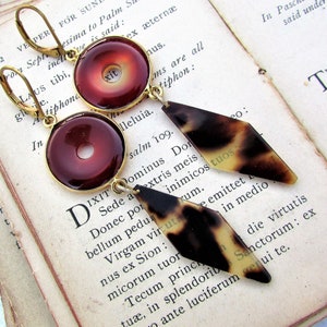 Puede incluir: Un par de pendientes colgantes con estética vintage. Cada pendiente presenta un colgante circular de color marrón rojizo con un marco dorado y un dije en forma de diamante con estampado de carey. Los pendientes se muestran sobre un fondo de páginas de libros antiguos.