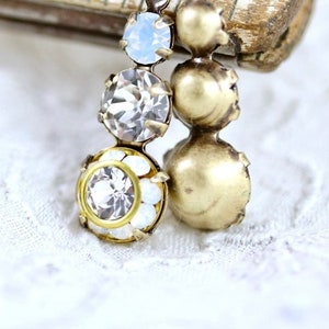 Puede incluir: Primer plano de pendientes de estilo vintage con tres cristales transparentes y un engaste de metal dorado. El cristal inferior está rodeado de pequeñas piedras opalinas blancas. Los pendientes están sobre un fondo de encaje blanco.