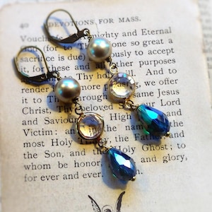 以下が含まれることがあります： ヴィンテージ感のあるドロップイヤリング。各イヤリングは、真珠のようなビーズ、透明なクリスタル、涙型の青緑色の宝石が特徴です。イヤリングは、古い本のページの背景に配置されています。