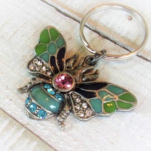 Könnte beinhalten: Ein dekorativer Schlüsselanhänger mit einem Schmetterlingsdesign. Der Schmetterling hat grüne, türkisfarbene und schwarze Emailleflügel, einen rosa Mittelstein und ist mit klaren Kristallen und blauen Steinen verziert. Der Schlüsselanhänger hat eine silberfarbene Metalloberfläche.