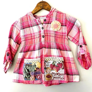 May include: Pink plaid long-sleeve shirt with a hood and two front pockets. The shirt features a floral applique and decorative patches with text and illustrations of strawberries and vegetables. The shirt has button closures and rolled-up sleeves.