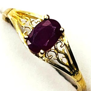 May include: A gold ring with an oval, deep red gemstone. The ring has an ornate filigree design on the sides of the band, with a polished finish. The gemstone is the focal point, set in a classic prong setting.