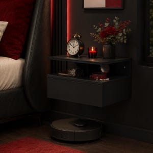 Puede incluir: Una mesita de noche flotante negra con un cajón y un estante abierto. La superficie superior sostiene un despertador analógico negro, una vela roja en un portavelas de cristal y un jarrón con rosas rojas y follaje. Un robot aspirador negro está debajo.