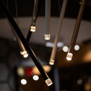 Op de afbeelding: Een moderne hanglamp met meerdere slanke, cilindrische staven in zwarte, witte en metallic afwerkingen. Elke staaf hangt verticaal en eindigt aan de onderkant in een kleine, heldere, oplichtende LED-lichtbron.