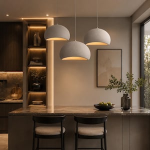 Puede incluir: Un conjunto de tres lámparas colgantes en forma de cúpula con un acabado gris claro mate, suspendidas del techo por cables blancos delgados. Cada luminaria presenta una pantalla ancha y curva que dirige luz cálida hacia abajo.