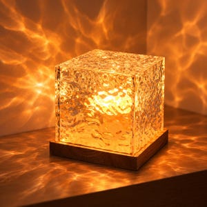 Peut inclure: Une lampe de table carrée dotée d'un abat-jour en cube de verre texturé et ondulé qui émet une lumière dorée chaude. Le cube de verre repose sur une base en bois carrée et plate, projetant des motifs lumineux complexes rappelant l'eau.