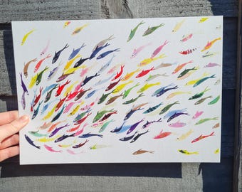 Könnte beinhalten: Ein rechteckiger Kunstdruck mit einem Schwarm bunter Fische. Die Fische sind in verschiedenen Farben dargestellt, darunter Gelb, Rot, Blau und Grün, vor einem weißen Hintergrund. Das Kunstwerk wird vor einer verwitterten Holzoberfläche gehalten.
