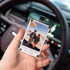 May include: A clear acrylic photo holder with a photo of a couple leaning against a vintage car at a gas station. The photo holder has a silver clip and the text "My Heart Rides Here. Drive Safe."