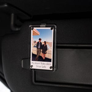 May include: Clear acrylic car visor photo holder with a photograph of a couple standing by a vintage car at a gas station. The photo has the text "My Heart Rides Here. Drive Safe."