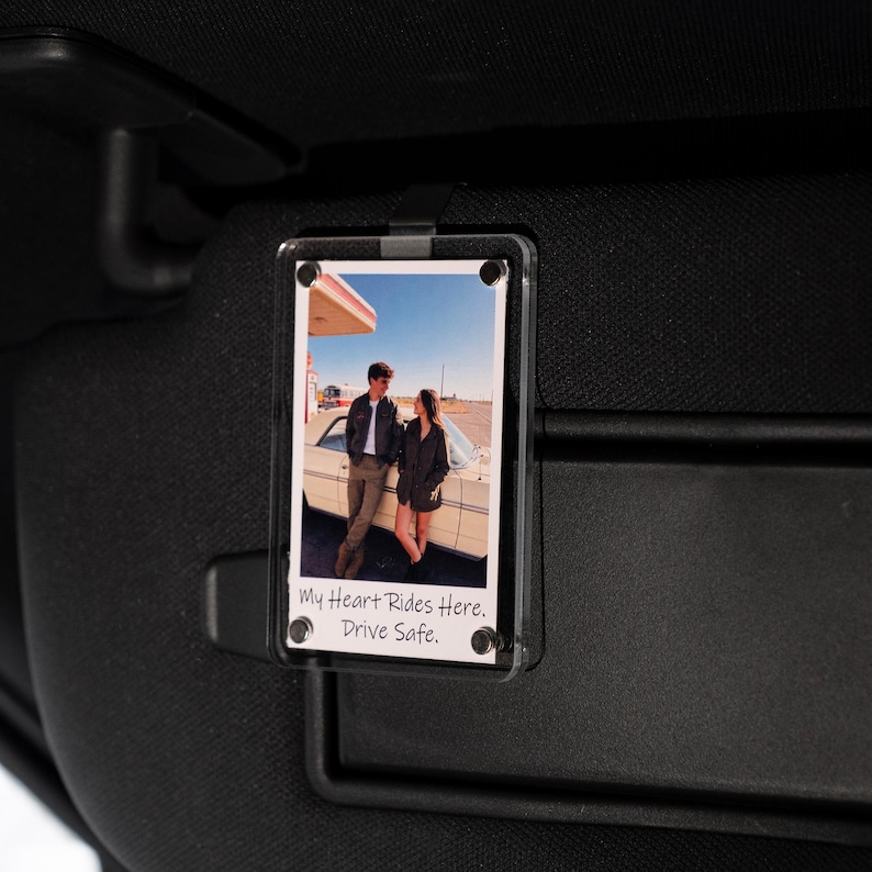 May include: Clear acrylic car visor photo holder with a photograph of a couple standing by a vintage car at a gas station. The photo has the text "My Heart Rides Here. Drive Safe."
