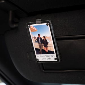May include: Clear acrylic car visor photo holder with a photograph of a couple standing near a vintage car. The photo holder is attached to a black car visor. Text on the photo reads: "My Heart Rides Here. Drive Safe."