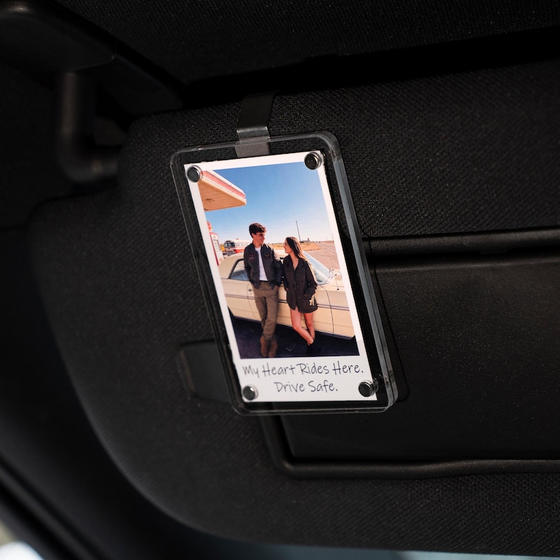 May include: Clear acrylic car visor photo holder with a photograph of a couple standing near a vintage car. The photo holder is attached to a black car visor. Text on the photo reads: "My Heart Rides Here. Drive Safe."