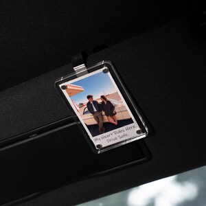 May include: Clear acrylic car visor photo holder with a picture of a couple. The photo holder is rectangular and has the text "My Heart Rides Here. Drive Safe." The visor is black.