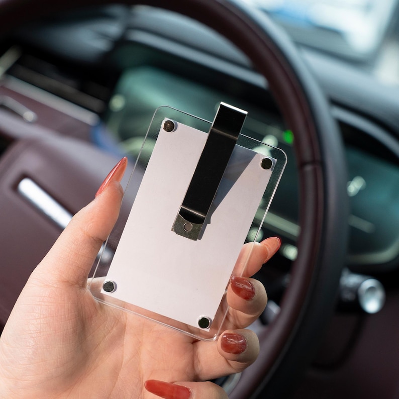 May include: A clear acrylic card holder with a black metal clip. The holder is rectangular with rounded corners and is held in front of a car's steering wheel. The clip is positioned vertically in the center.