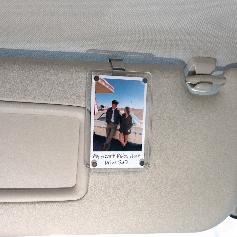 May include: A clear acrylic photo holder clipped to a car visor. The holder contains a vintage photograph of a couple posing by a car. The text "My Heart Rides Here. Drive Safe." is printed at the bottom of the photo.