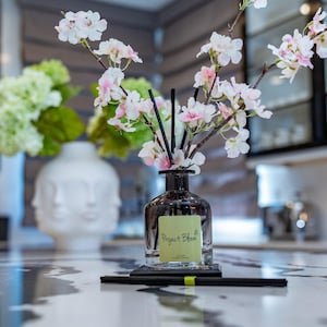 Pode incluir: Arranjo decorativo com um difusor de vidro transparente, varetas pretas e um rótulo que diz "Project Bloom". Contém flores brancas e rosas. Um vaso branco com rostos ao fundo. Sobre uma bancada.