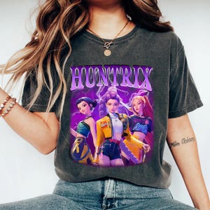 May include: Dark gray t-shirt featuring a graphic of three anime-style women with the word "HUNTRIX" in purple. The graphic has a purple lightning bolt background. The women are wearing colorful outfits.