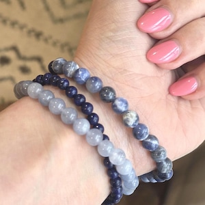 May include: Three beaded bracelets in shades of blue and gray. The bracelets are made of round beads and are stacked on a wrist. The nails are painted a light pink color.