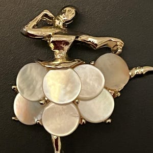 May include: A gold-toned ballerina brooch with a skirt made of round, iridescent mother-of-pearl discs. The dancer is posed mid-leap, arms extended. The brooch is set against a dark background.