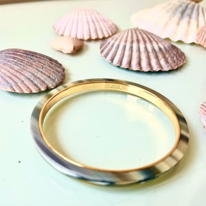 Op de afbeelding: Een goudkleurige armband met een gemarmerde, grijs-crèmekleurige band. De armband is gemarkeerd met de merknaam "J.CREW". Schelpen liggen verspreid rond de armband op een lichtgroen oppervlak.