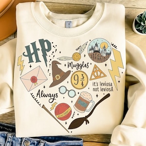 Pode incluir: Camisola creme com um gráfico em forma de coração com elementos de uma série de livros de fantasia. O design inclui um raio, um chapéu de bruxo, óculos e o texto "Always".